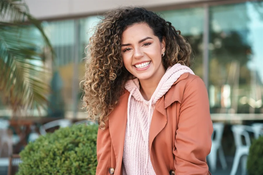 A smiling young woman wearing a tan jacket and jeans showcasing a stunning smile makeover.